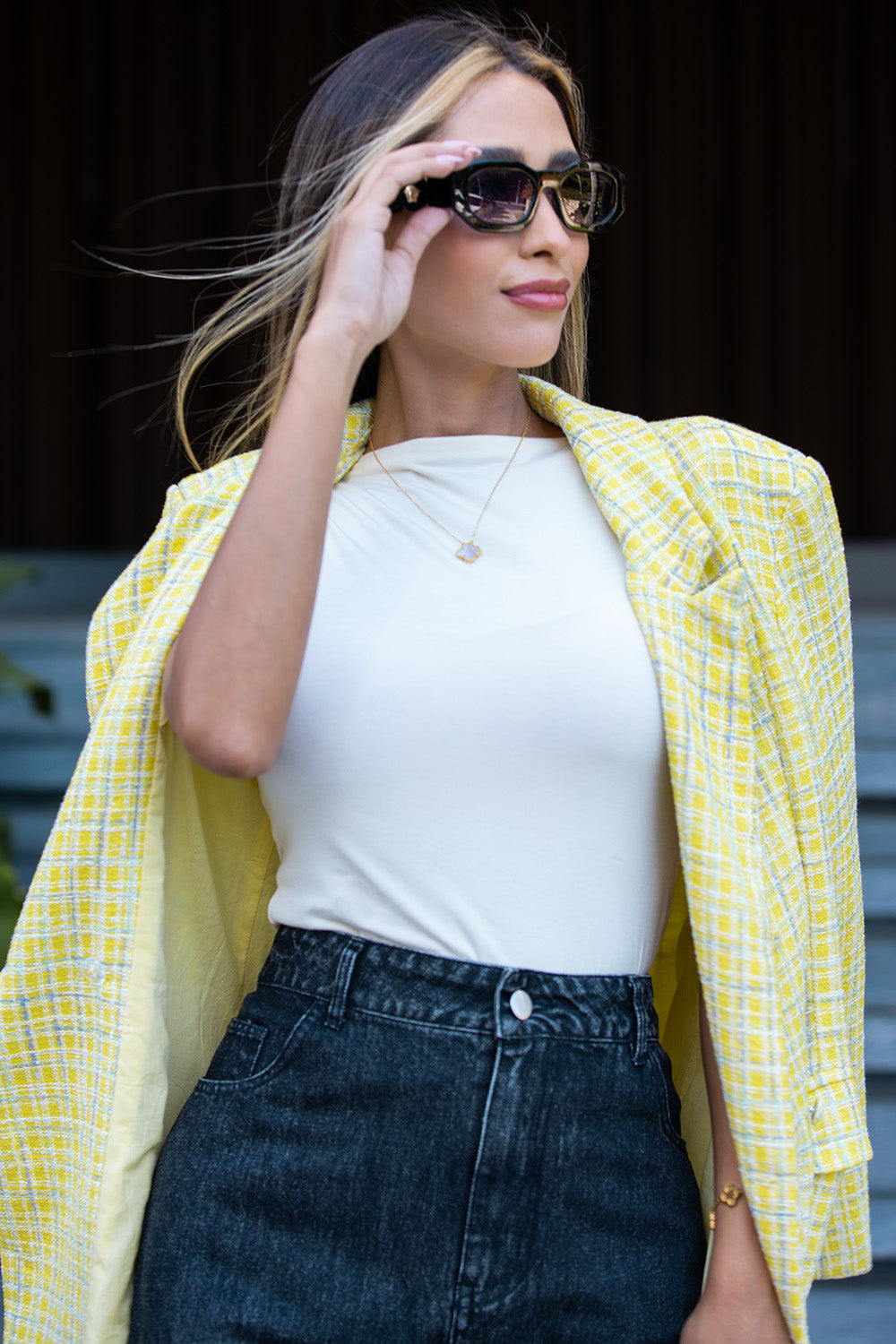 Woman wearing a yellow checkered jacket over a white top and dark jeans, adjusting sunglasses. Fine Modesty