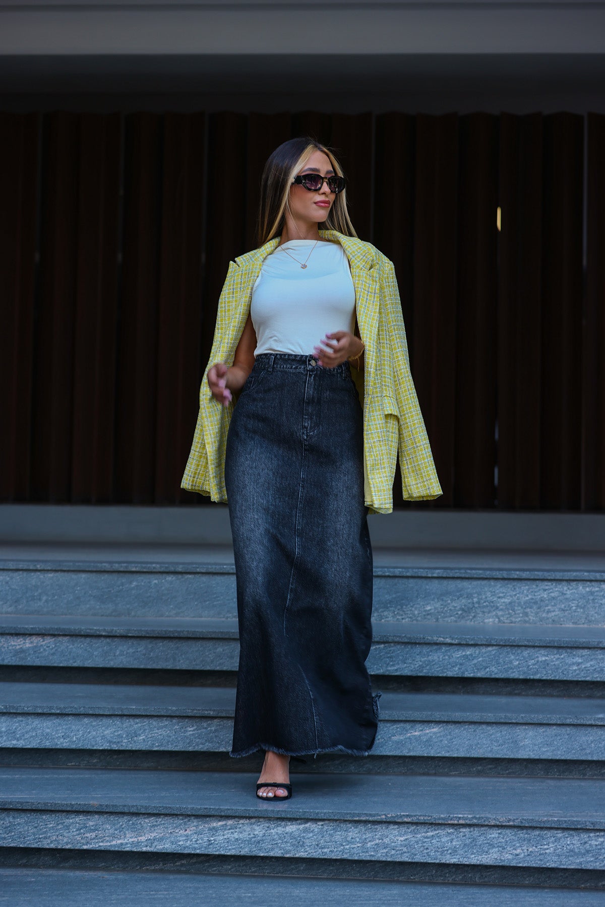 Woman in a yellow jacket and black skirt standing on steps with a dark background Fine Modesty